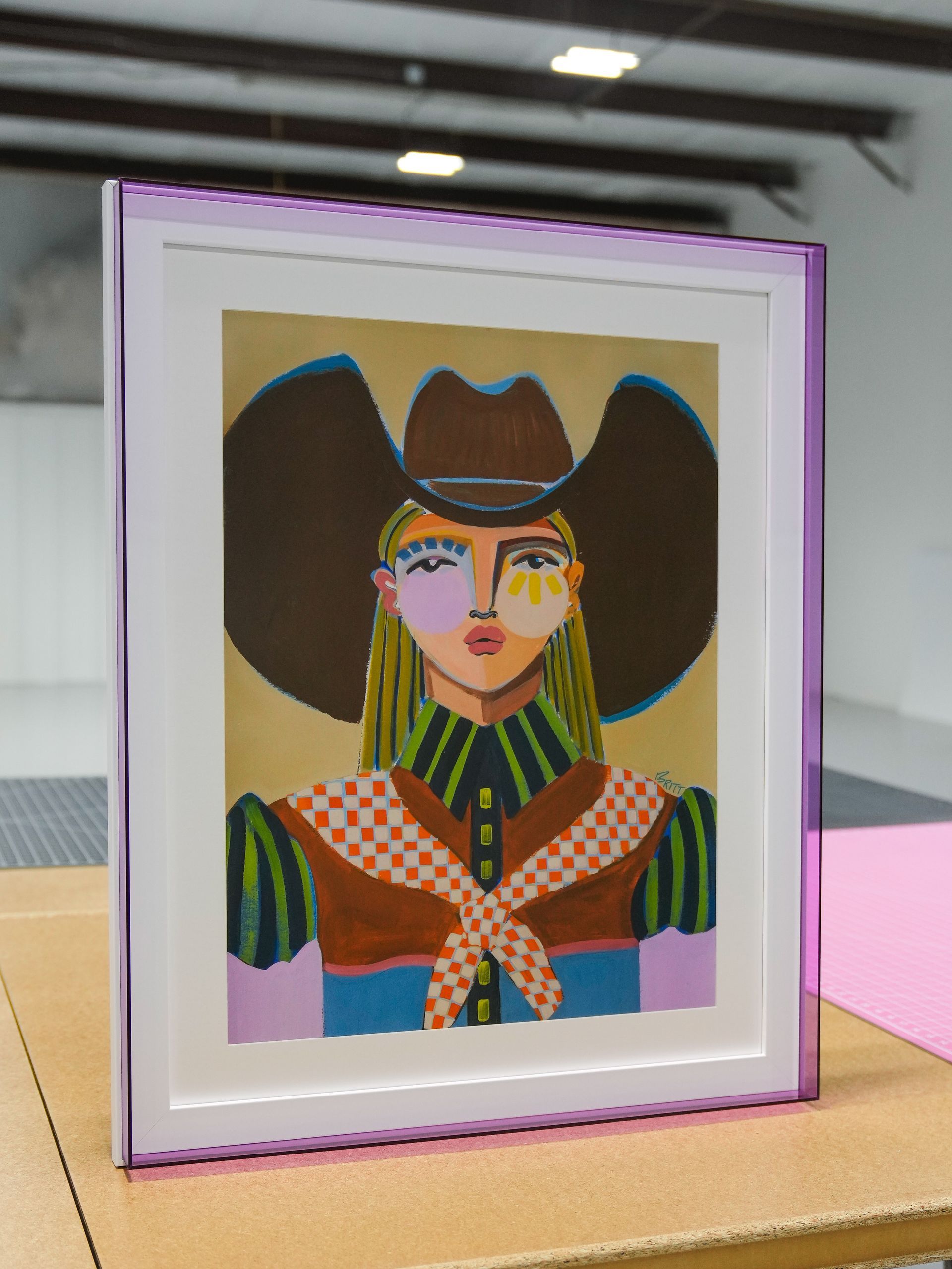 Framed colorful portrait illustration with a large hat, displayed on a table behind glass.