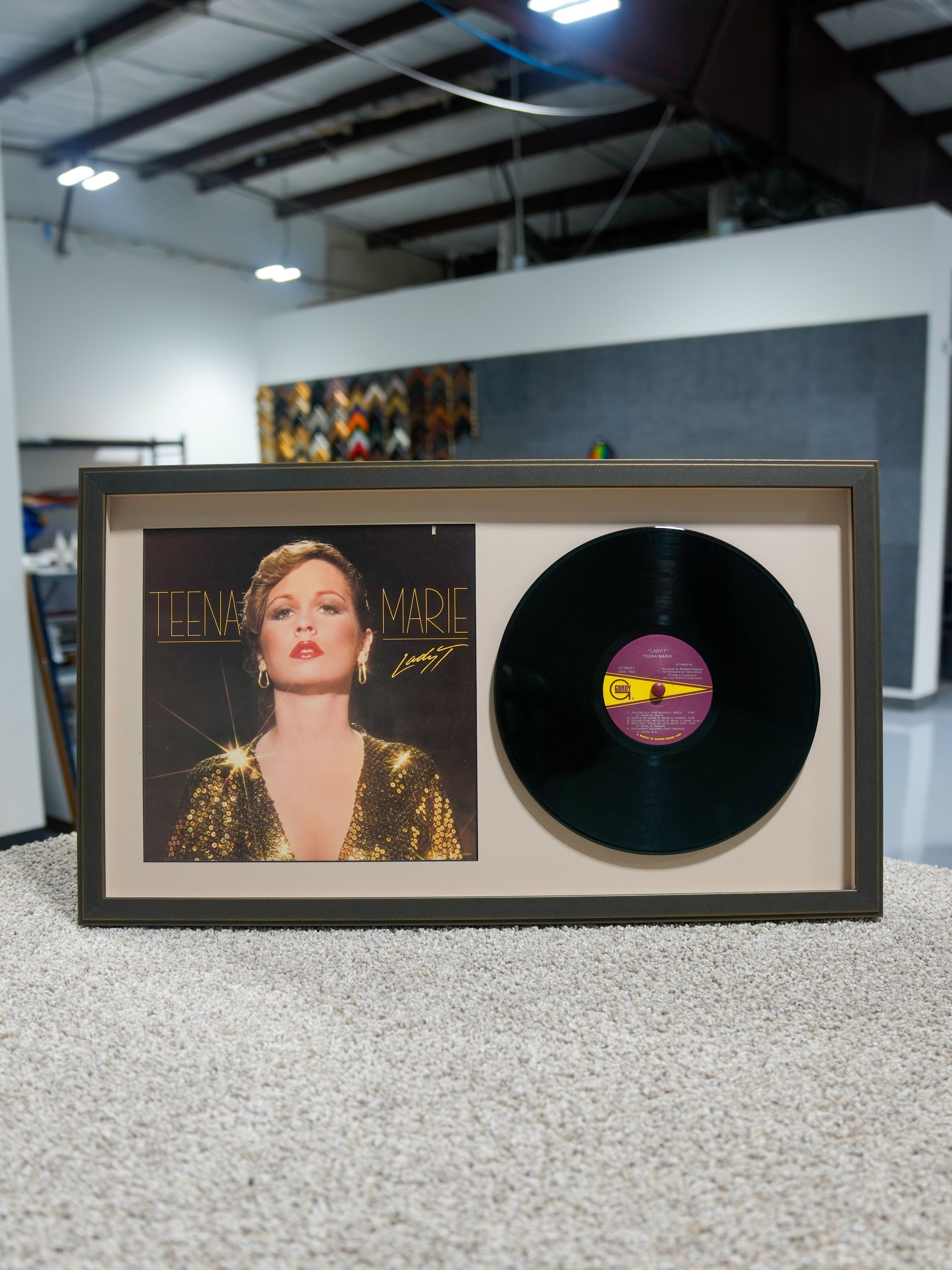 Framed vinyl record display with album cover and black disc on a carpeted floor in a modern room