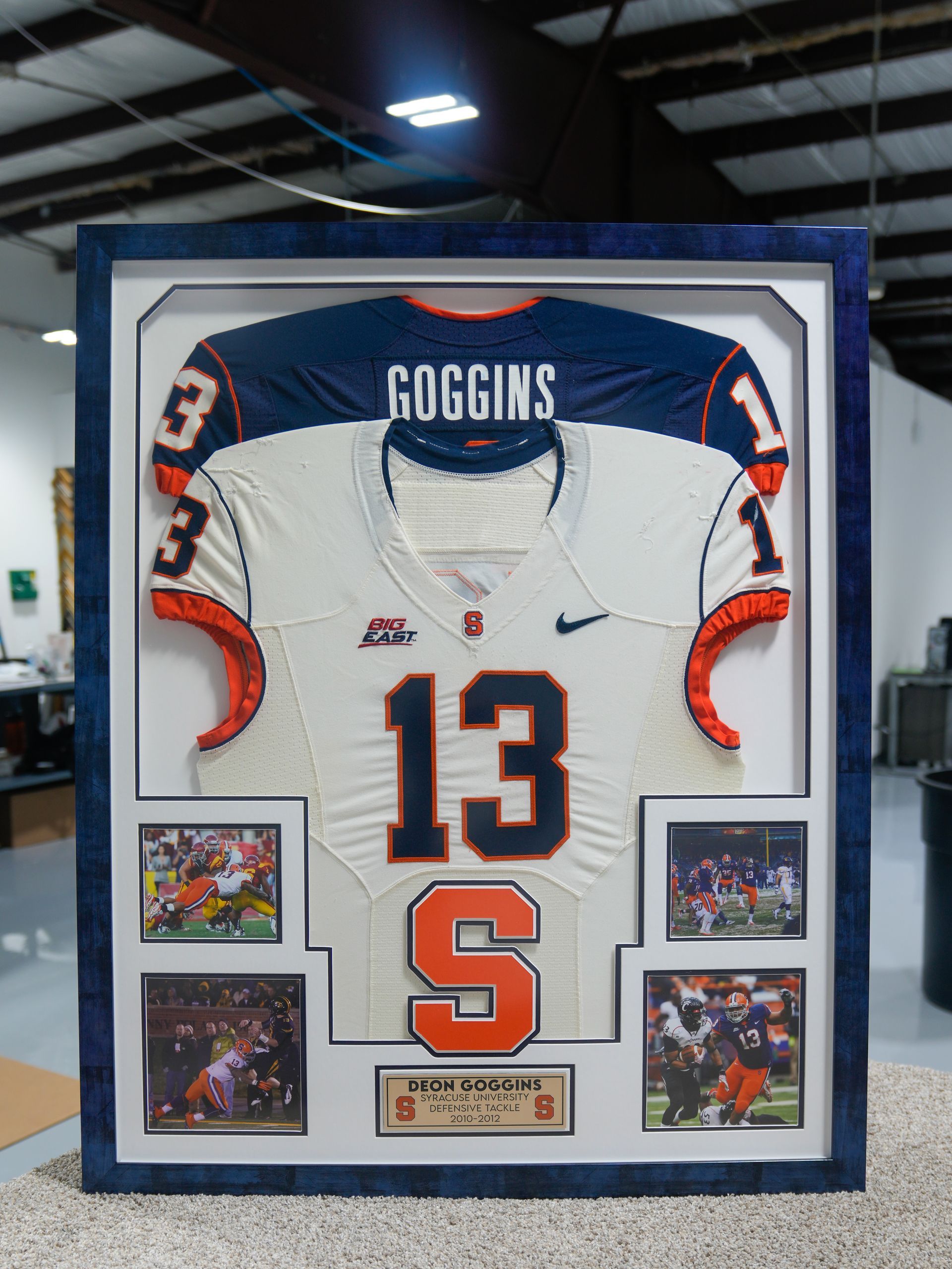 Framed white Syracuse football jersey with orange number 13 and Goggins name, displayed in a gallery.