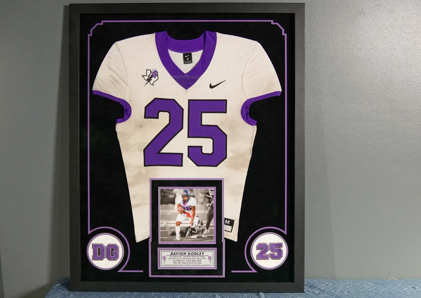 Framed white and purple football jersey numbered 25 with a small plaque below, displayed on a wall.