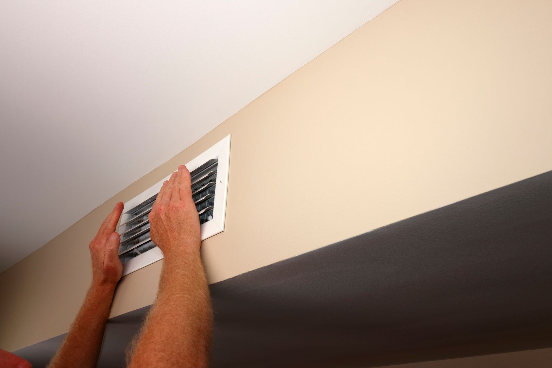 Hands installing a white vent cover on a beige wall near the ceiling.
