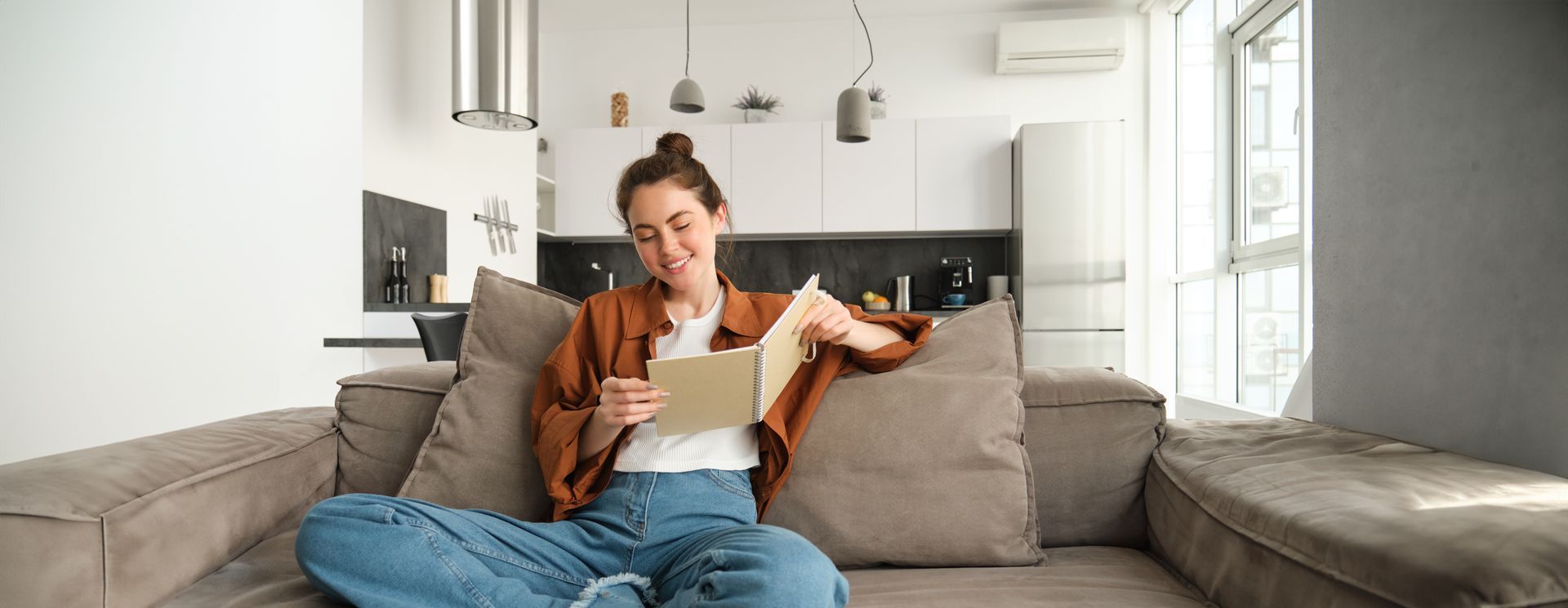 Person relaxing on a gray couch, reading a book in a bright modern living room.