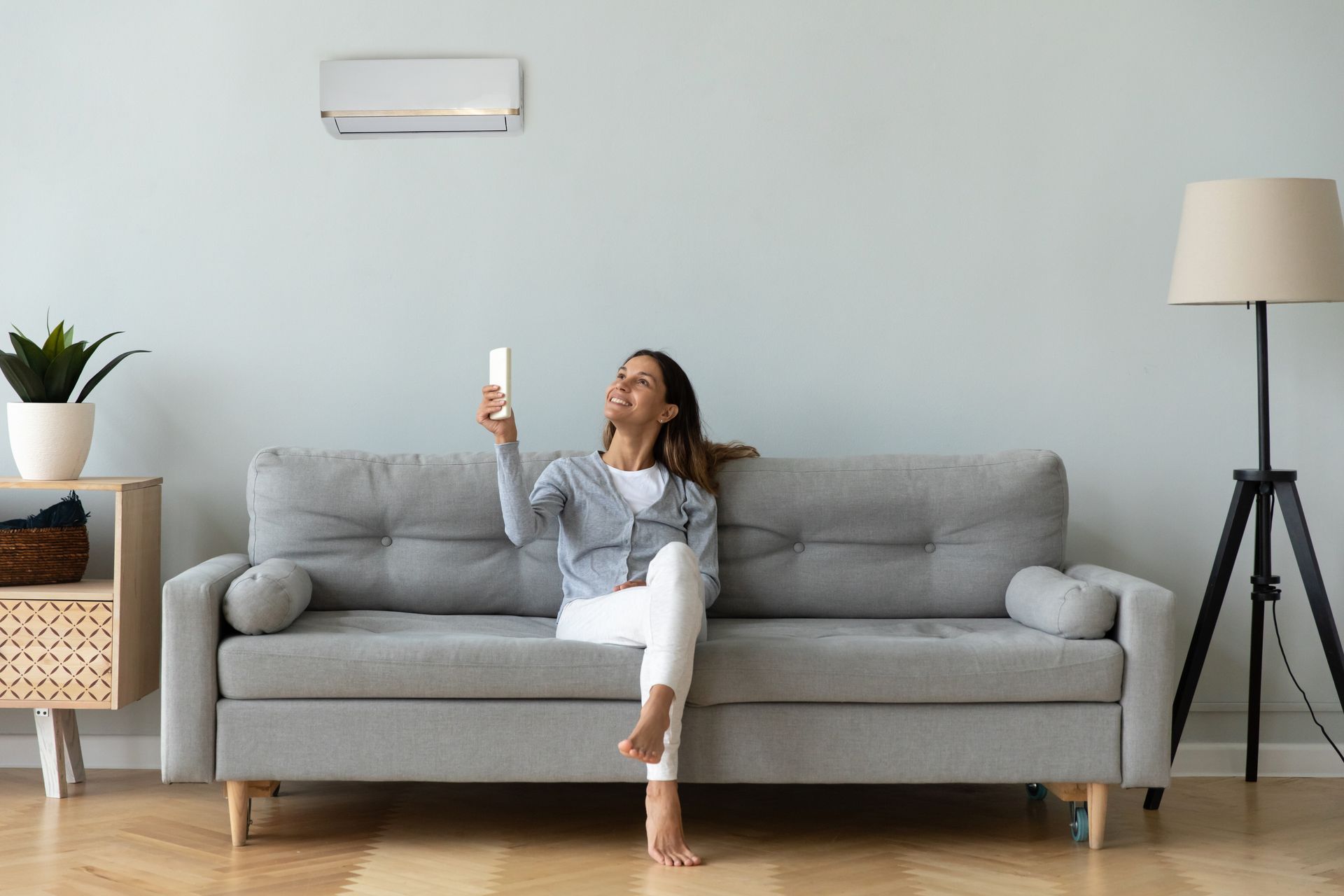 Woman sitting on a gray sofa, holding a phone in a bright living room with a floor lamp and plant.