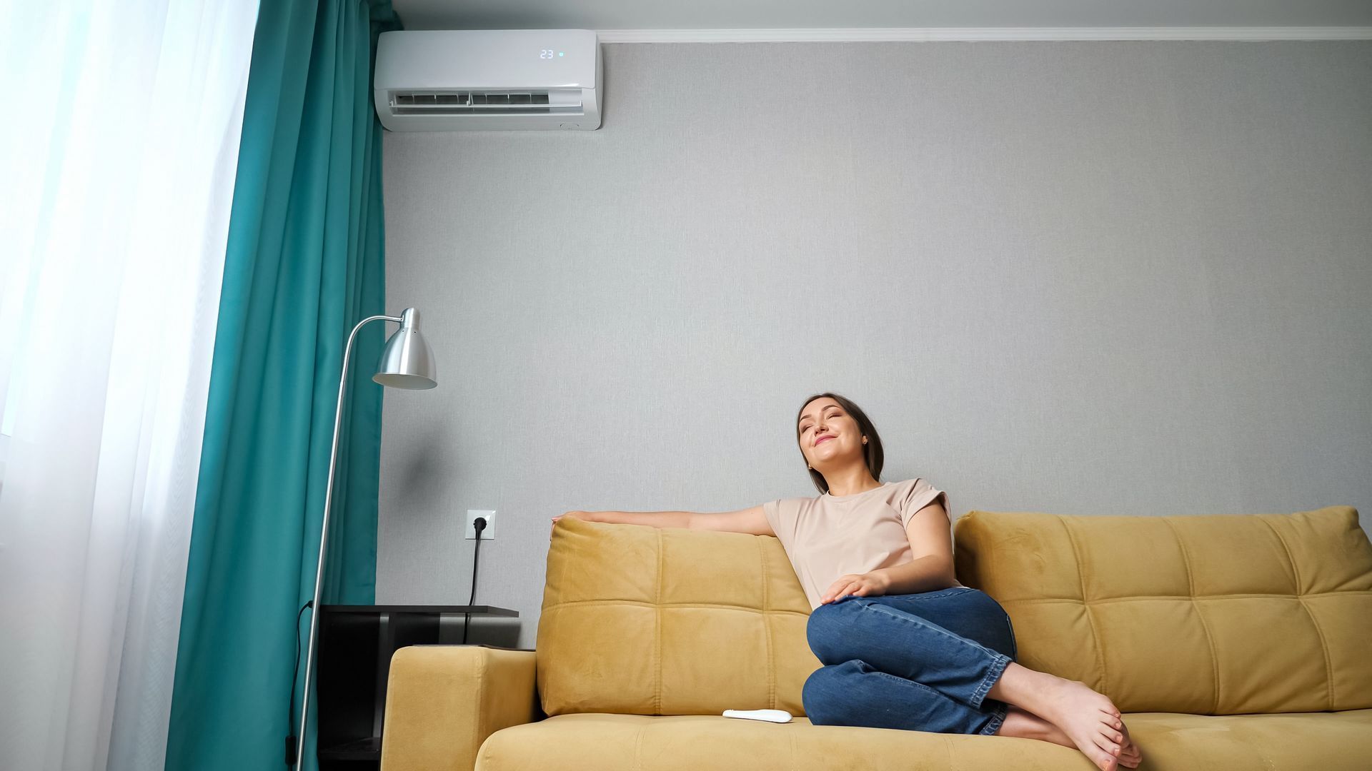 Person lounging on a yellow sofa in a bright living room with teal curtains and a wall-mounted air conditioner