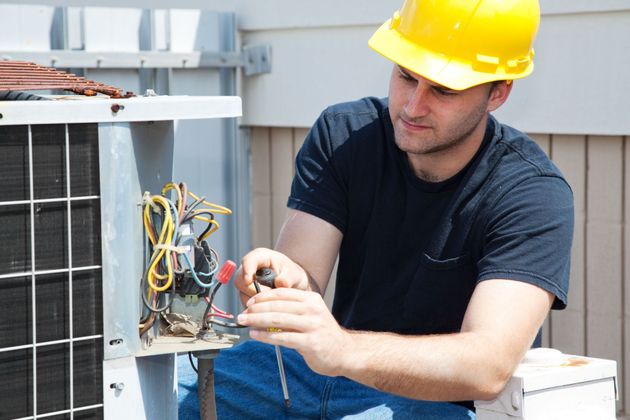 Technician in yellow hard hat repairing rooftop HVAC unit with exposed wiring