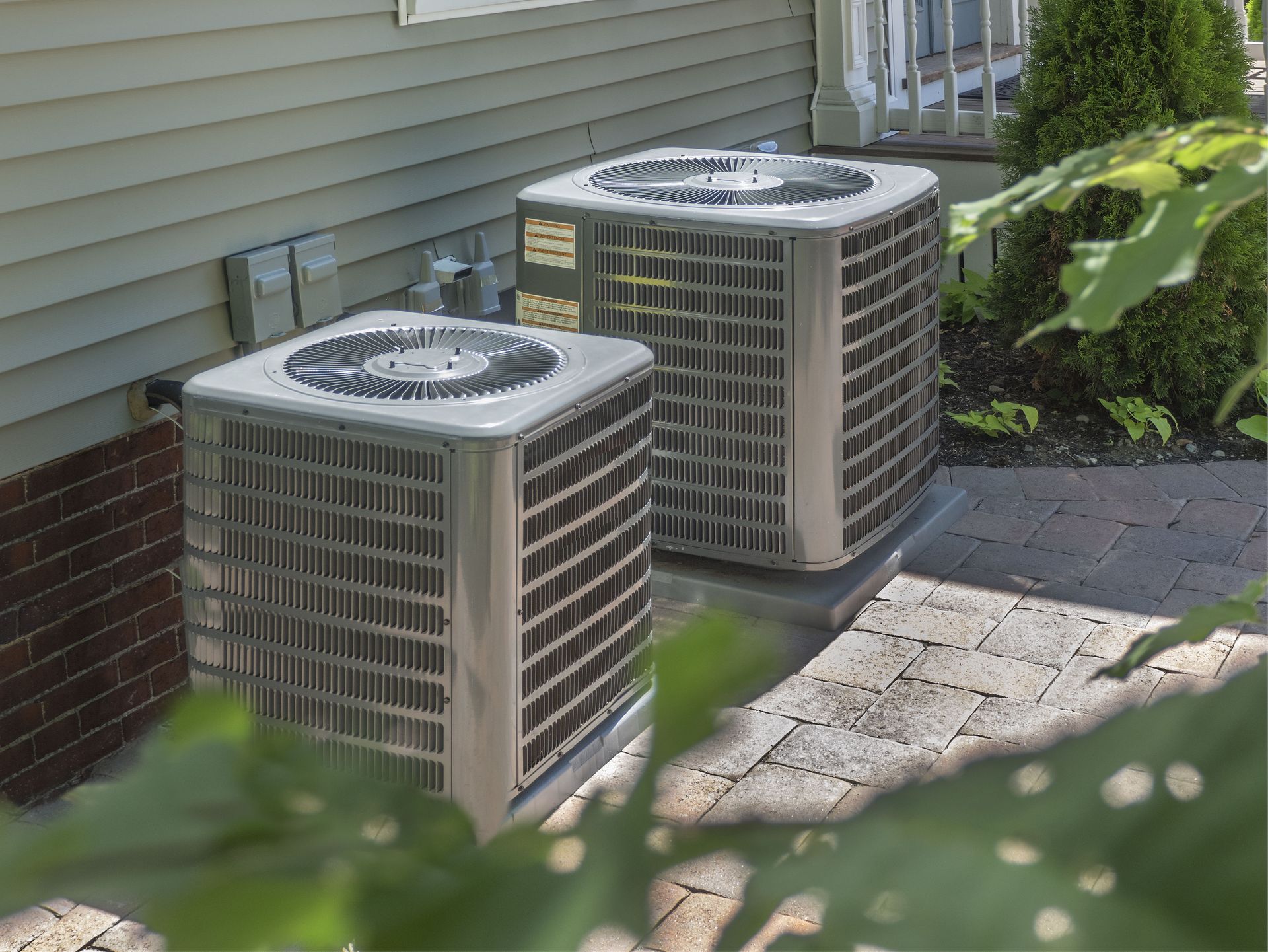Two outdoor air conditioning units beside a house on a paved patio, partially obscured by leaves