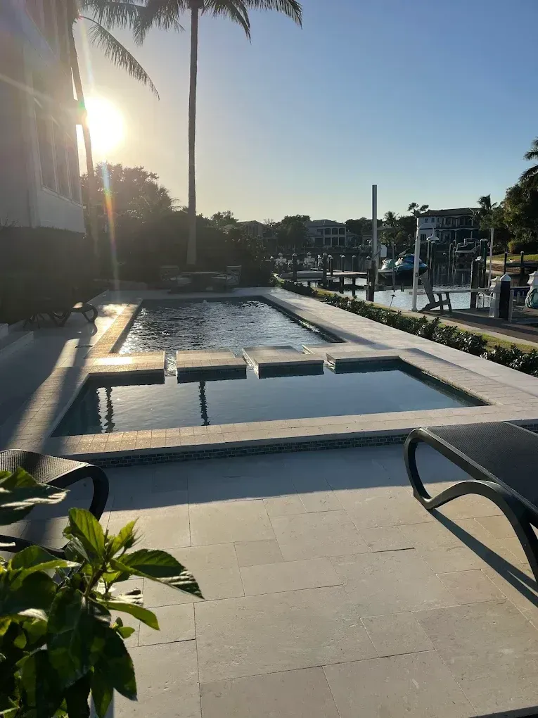 Poolside view at sunrise, rectangular pool with steps, lounge chairs, palm tree, harbor in the background.