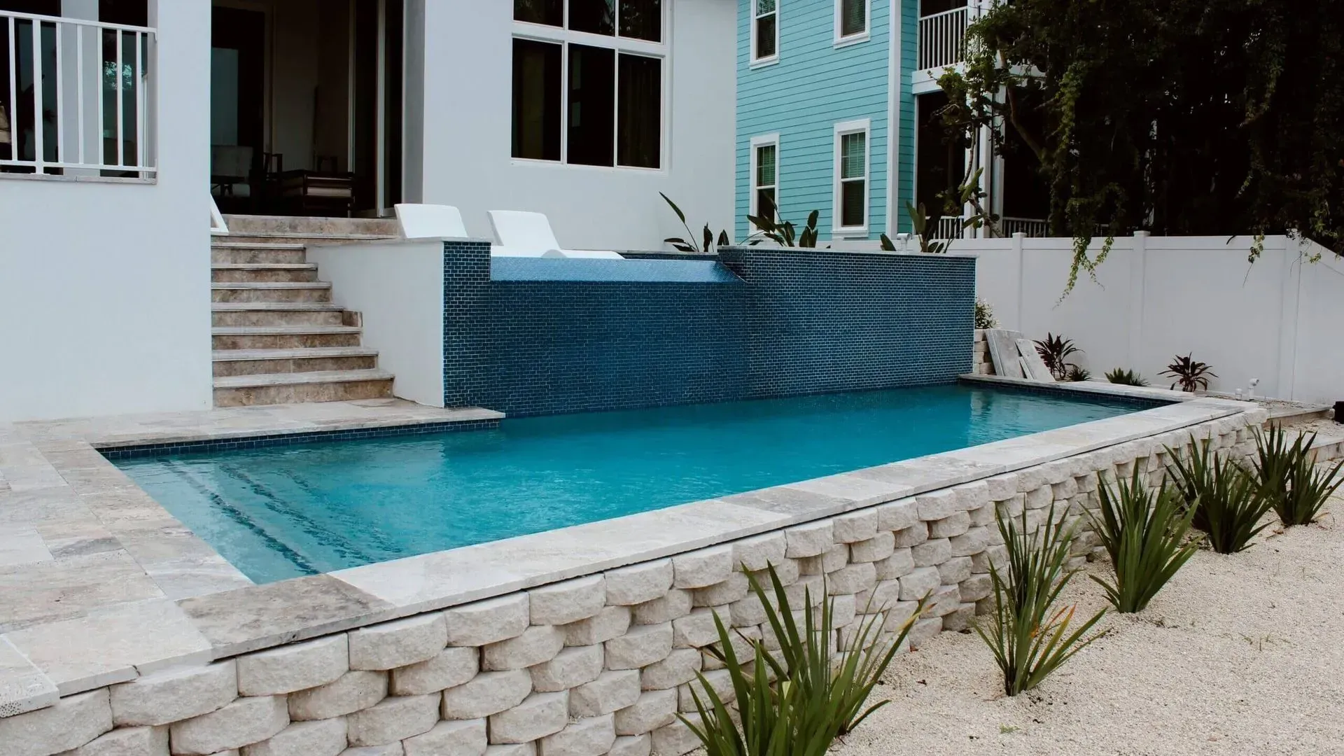 Outdoor swimming pool with blue tiles, steps, and plants; white building in background.