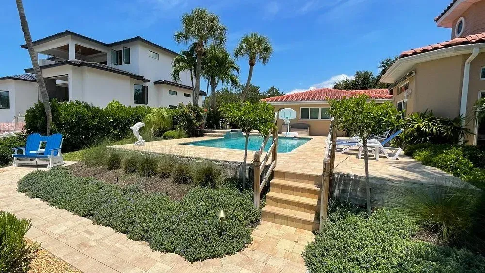 Backyard with pool, stone walkway, lush green landscaping, and houses under a blue sky.