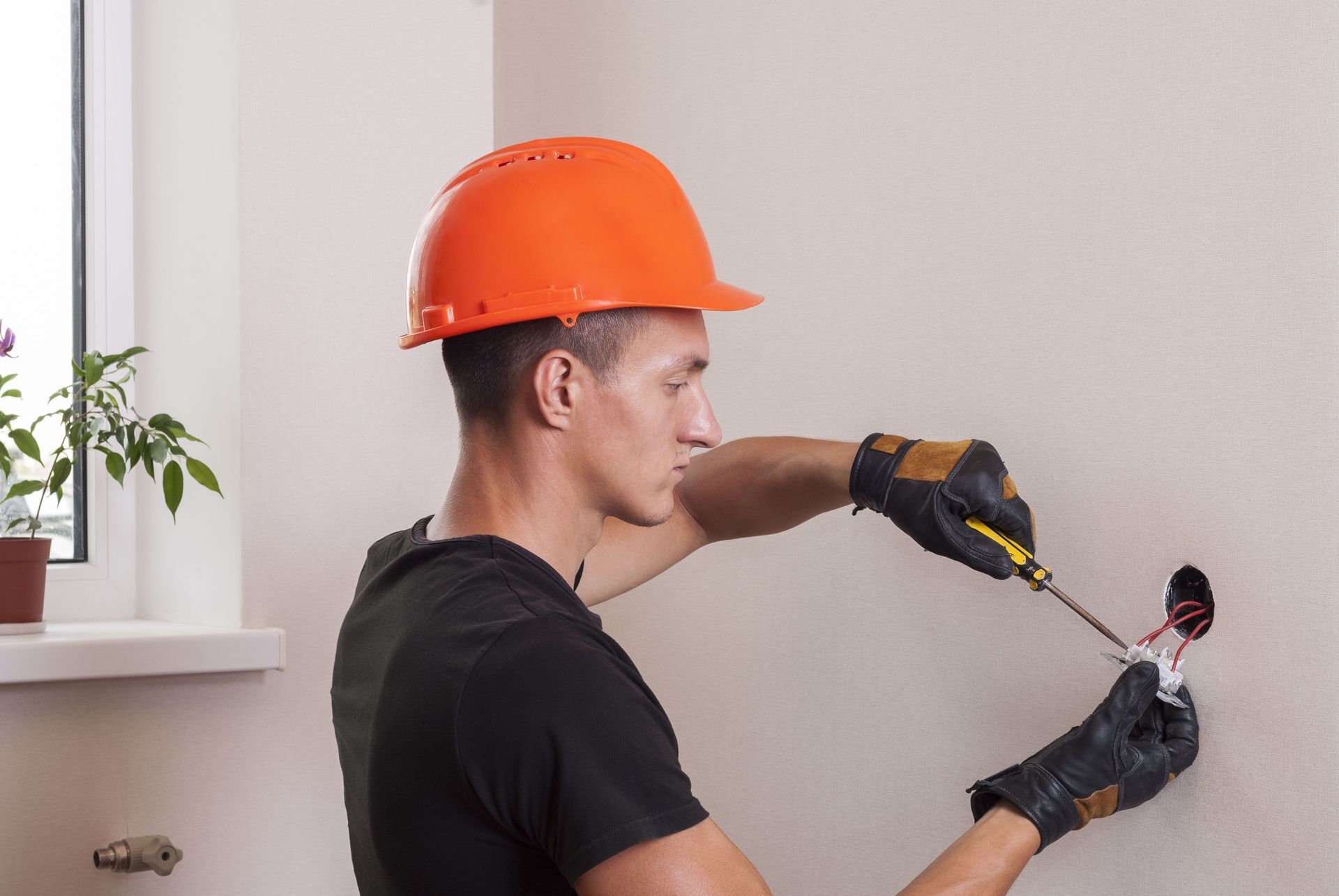 An electrician wearing a hard hat and protective gloves installs a light switch in a wall.