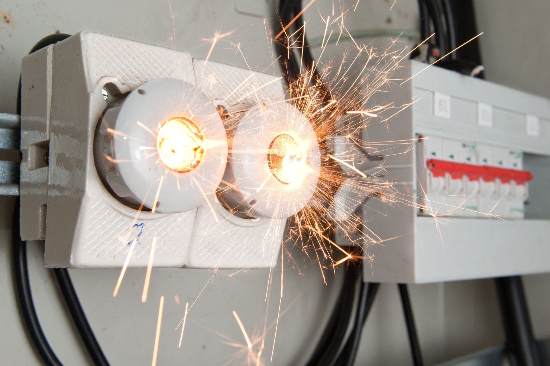 Sparks fly from a ceramic electrical fuse block mounted on a wall next to a circuit breaker panel.