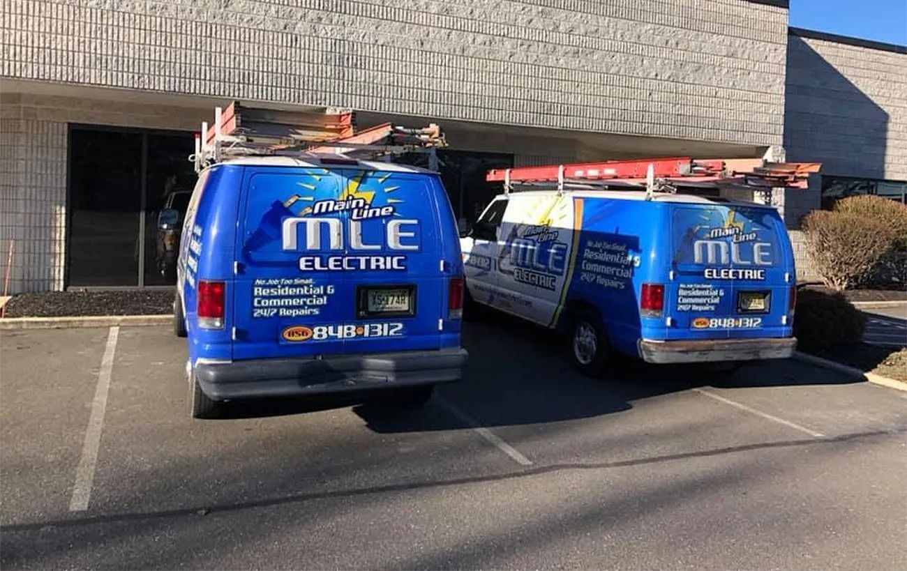 Two blue MLE Electric vans with ladders on their roofs parked in front of a commercial building.