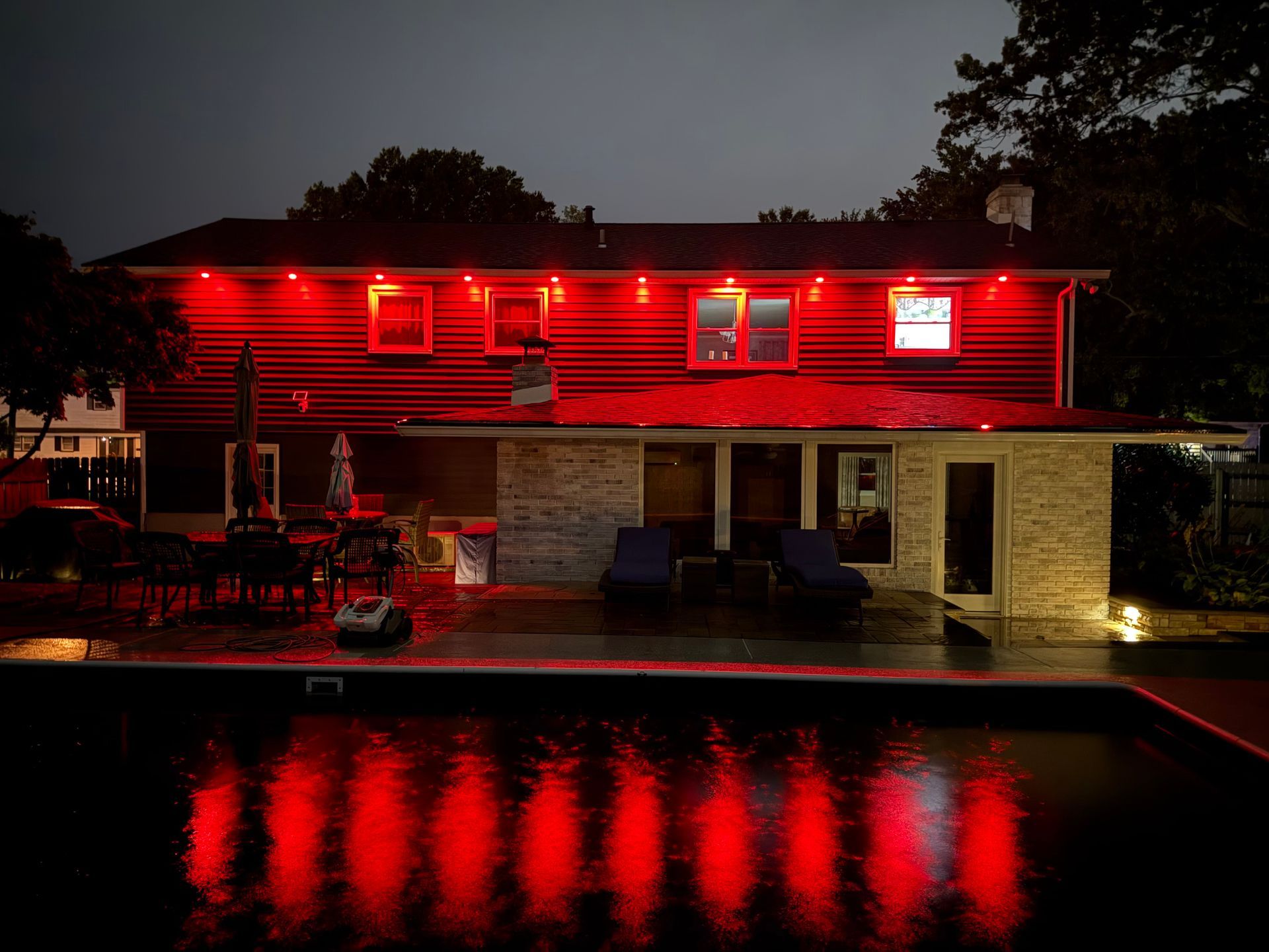 A two-story house at night illuminated by red exterior lights, with their vibrant reflections shimmering on a pool surface.