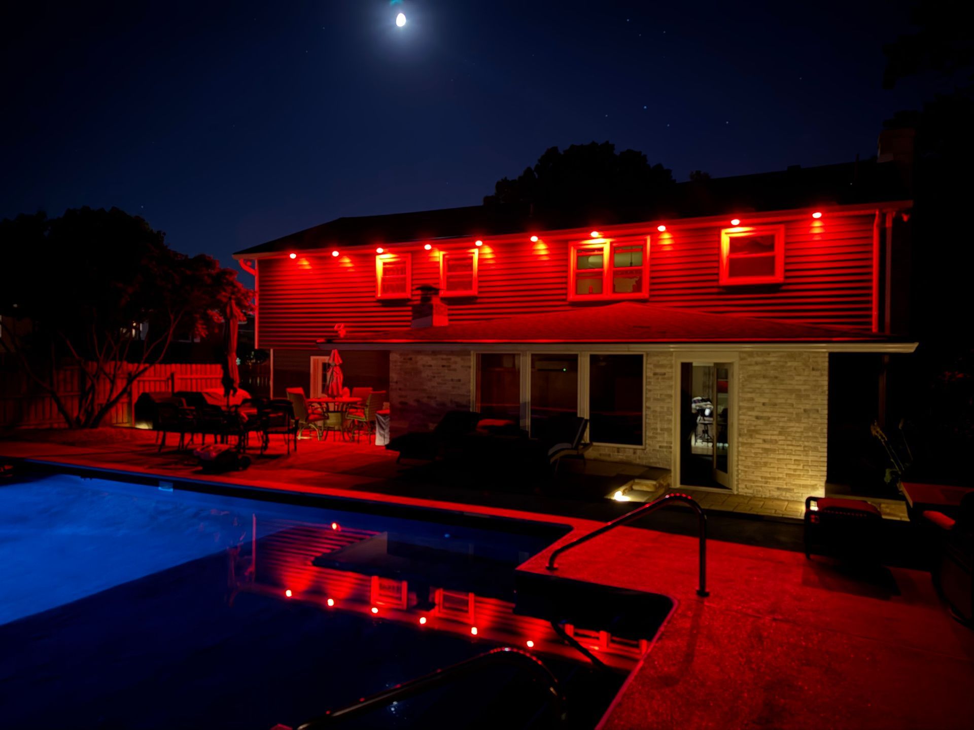 A two-story house at night illuminated by bright red outdoor lights, next to a pool reflecting the glow under a full moon.