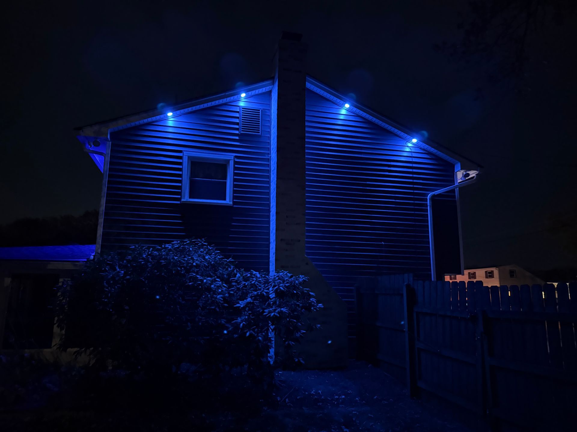 A dark exterior house wall at night, illuminated by four blue accent lights mounted under the roof eaves.