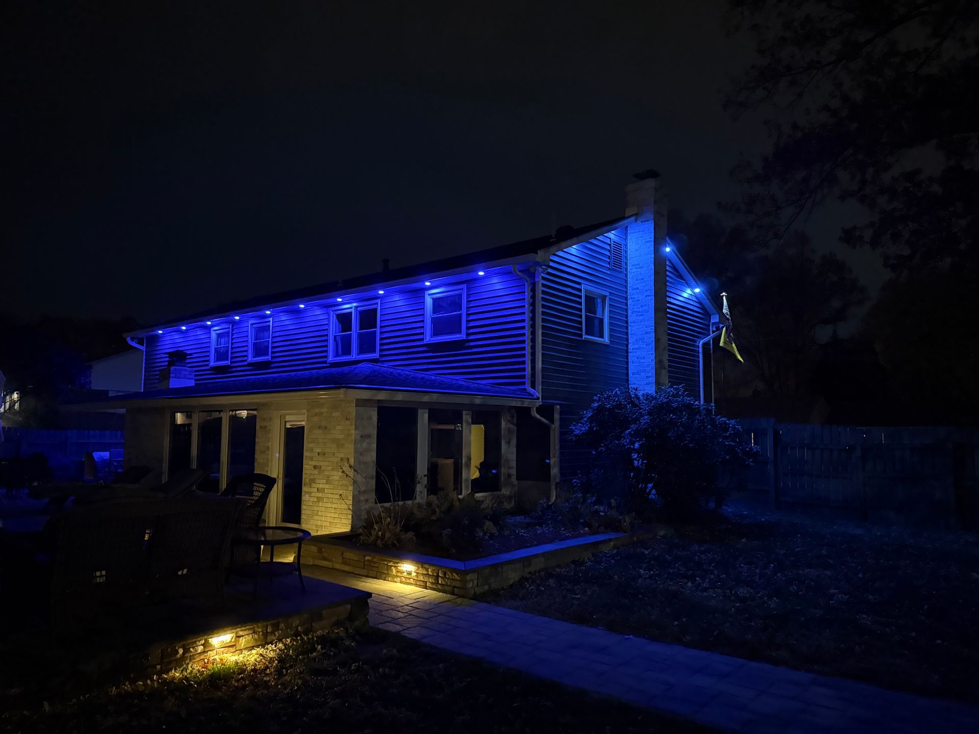 A residential house exterior at night illuminated by bright blue roofline accent lights and warm yellow ground spotlights.