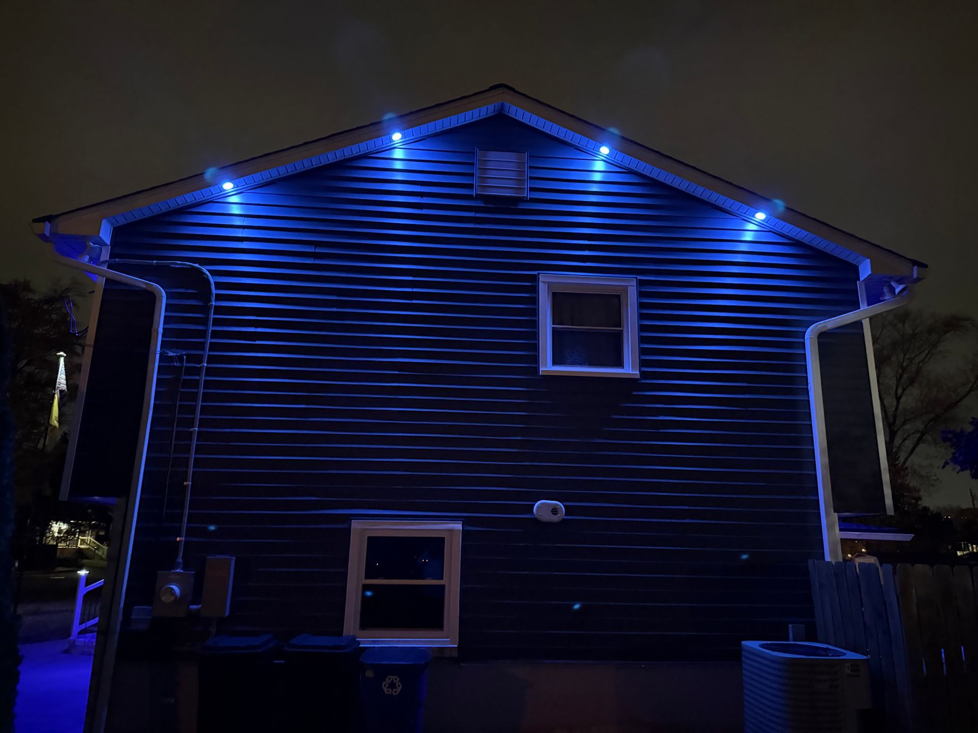 Dark blue exterior wall of a house at night, illuminated by four blue LED accent lights tucked under the roofline.