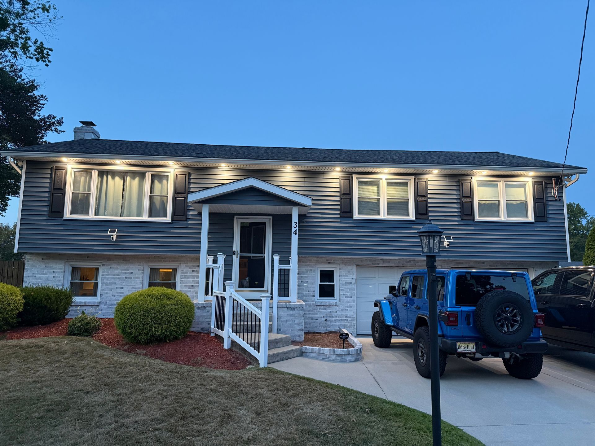 A blue split-level house with white stone trim, exterior lights, and a blue Jeep parked in the driveway at dusk.