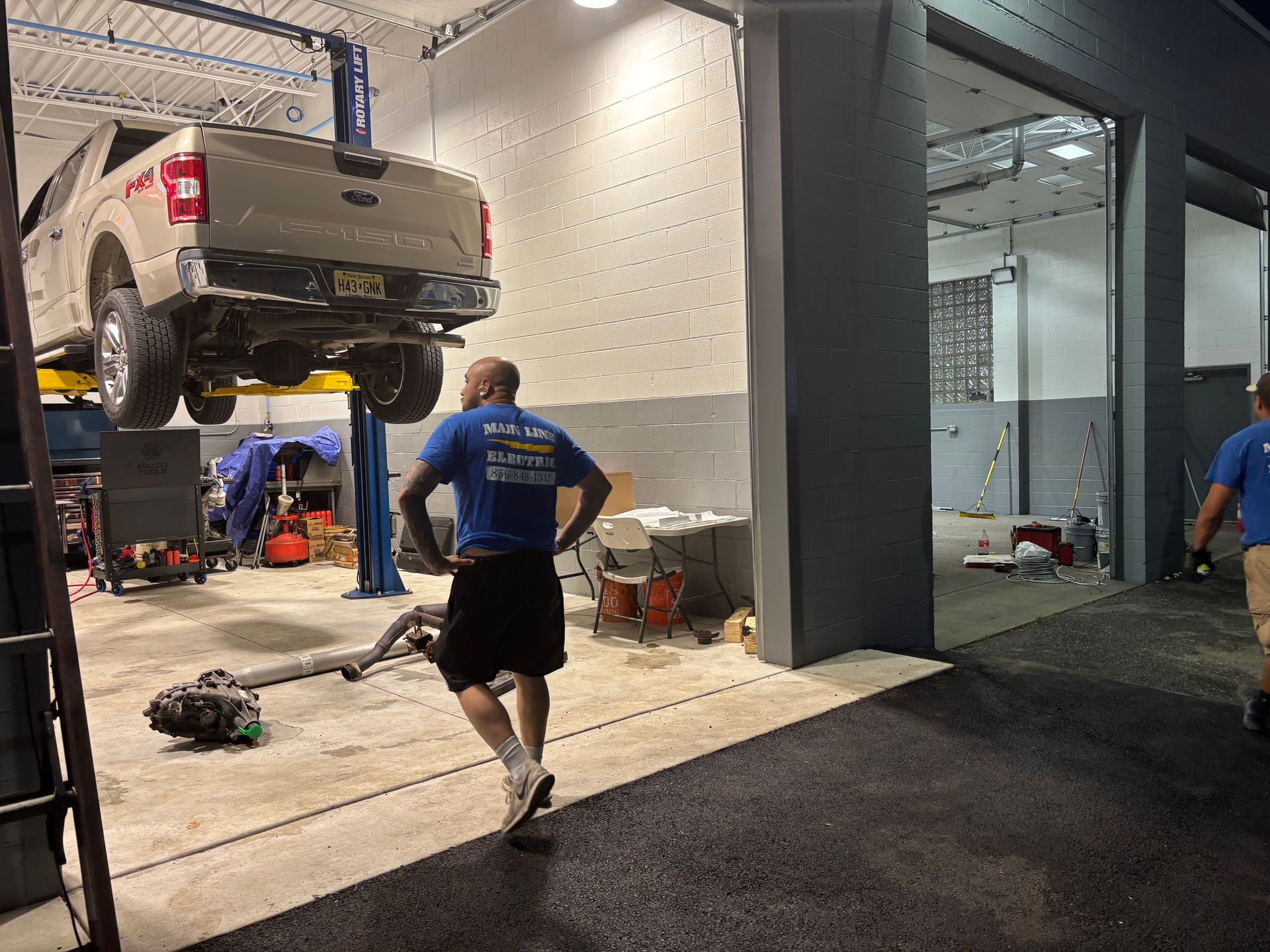 A mechanic in a blue shirt looks at a light-colored truck on a lift inside a vehicle repair garage.