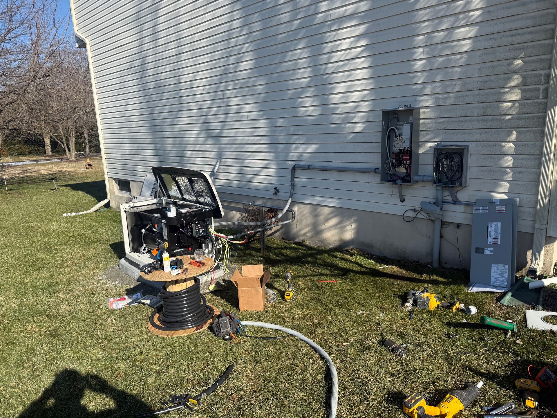 A standby generator under installation outside a house, with exposed electrical boxes on the siding and tools nearby.