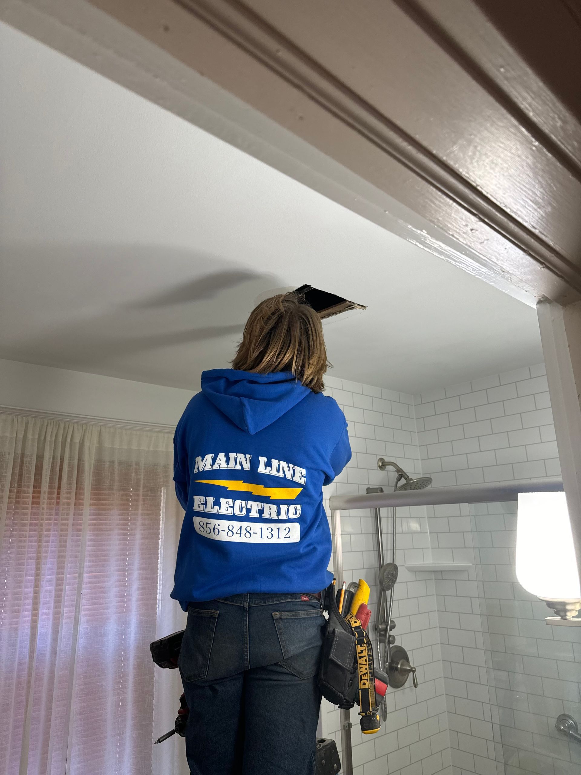 A worker in a blue hoodie stands on a ladder, reaching into an opening in a residential bathroom ceiling.