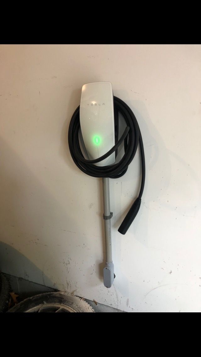 A white Tesla Wall Connector mounted on a white wall, displaying a glowing green status light with a coiled charging cable.