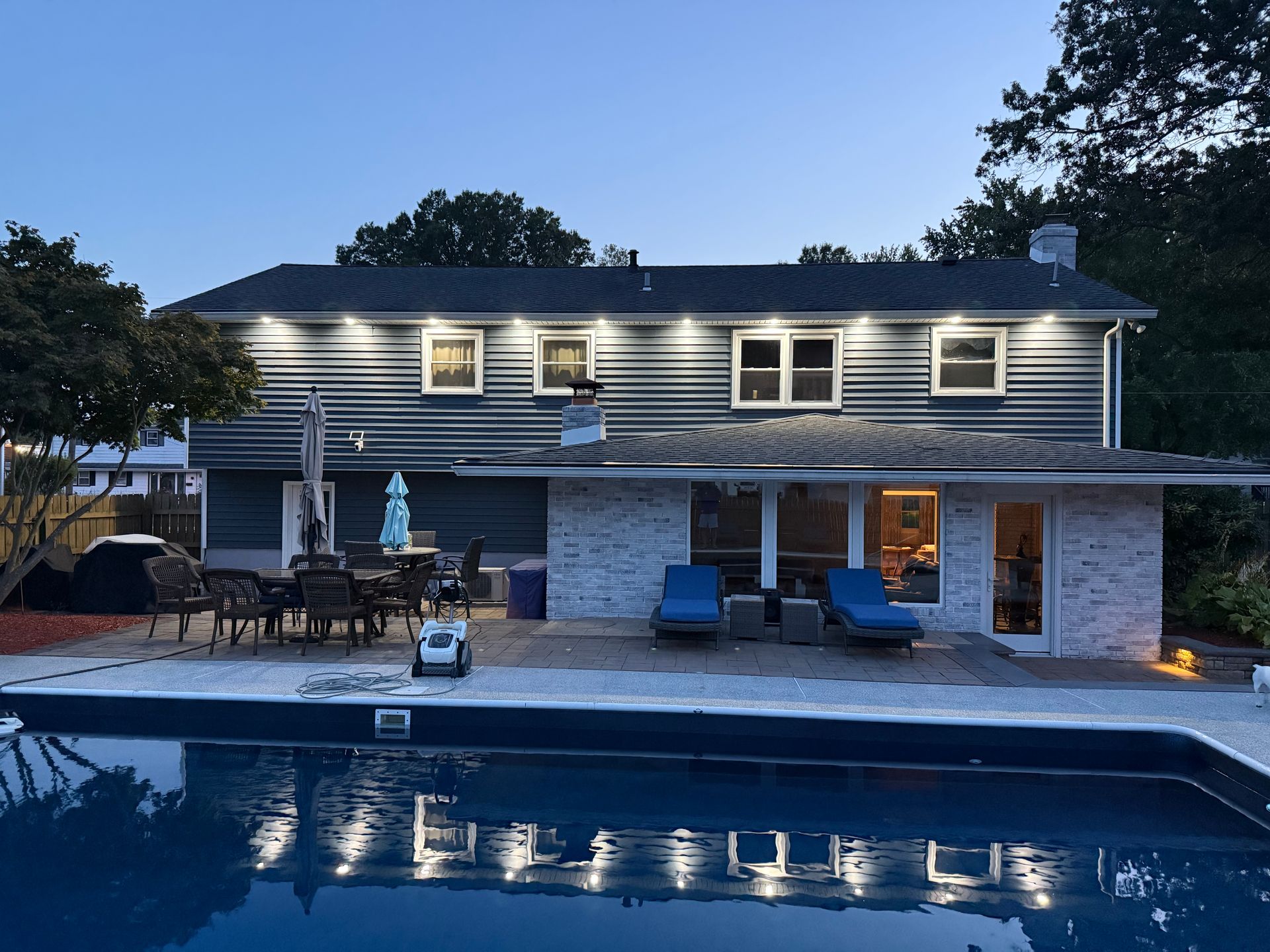 A two-story house at dusk with light reflecting on a backyard pool, patio furniture, and two lounge chairs.