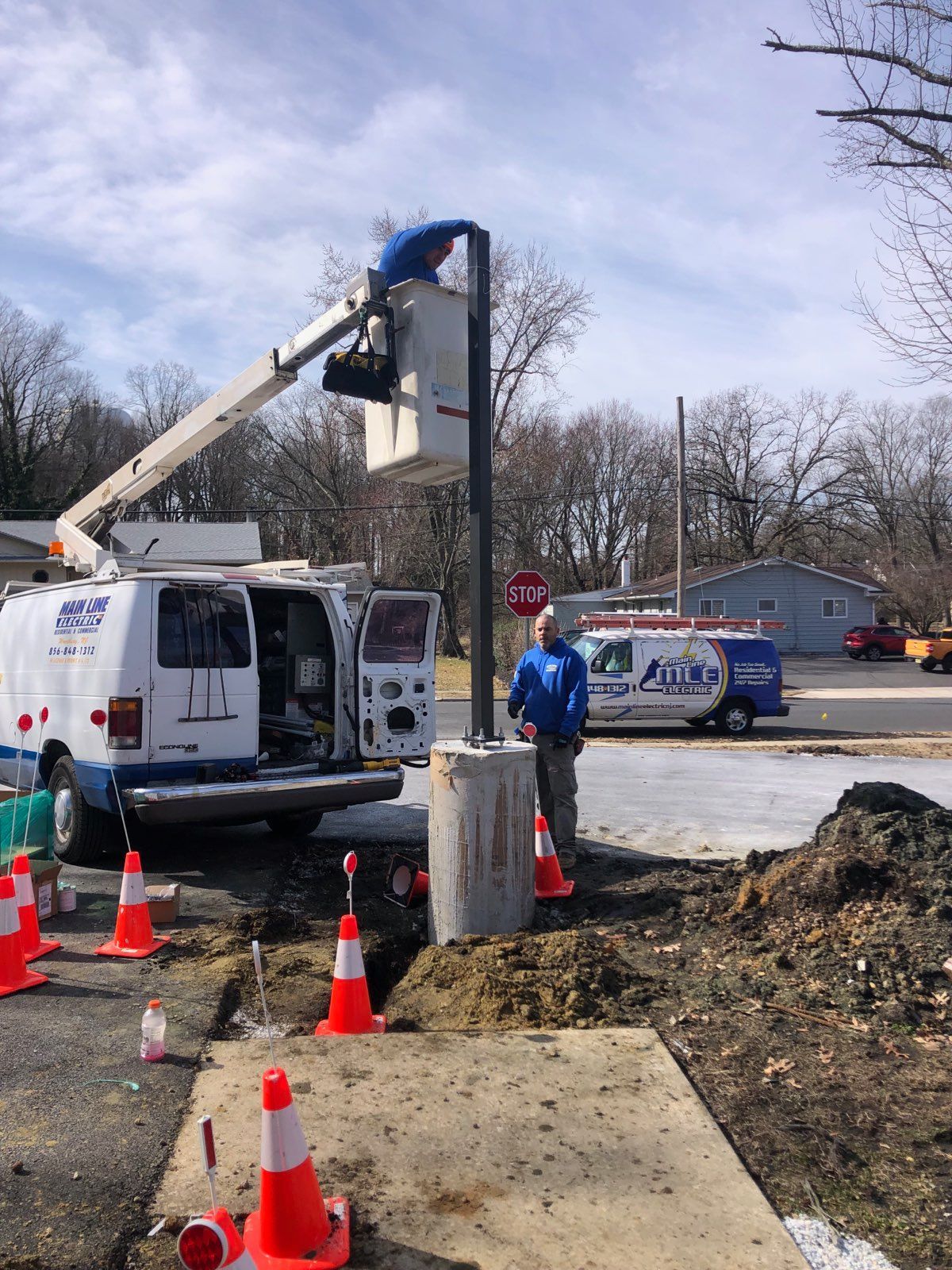 A worker in a bucket truck installs a tall metal pole on a concrete base with a colleague standing nearby on the ground.