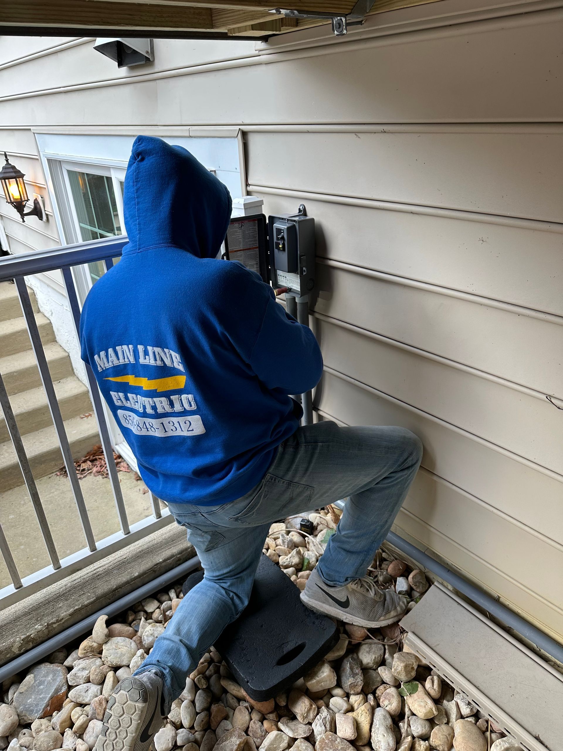 A person in a blue hoodie kneels on rocks, working on an electrical box mounted to the exterior siding of a house.