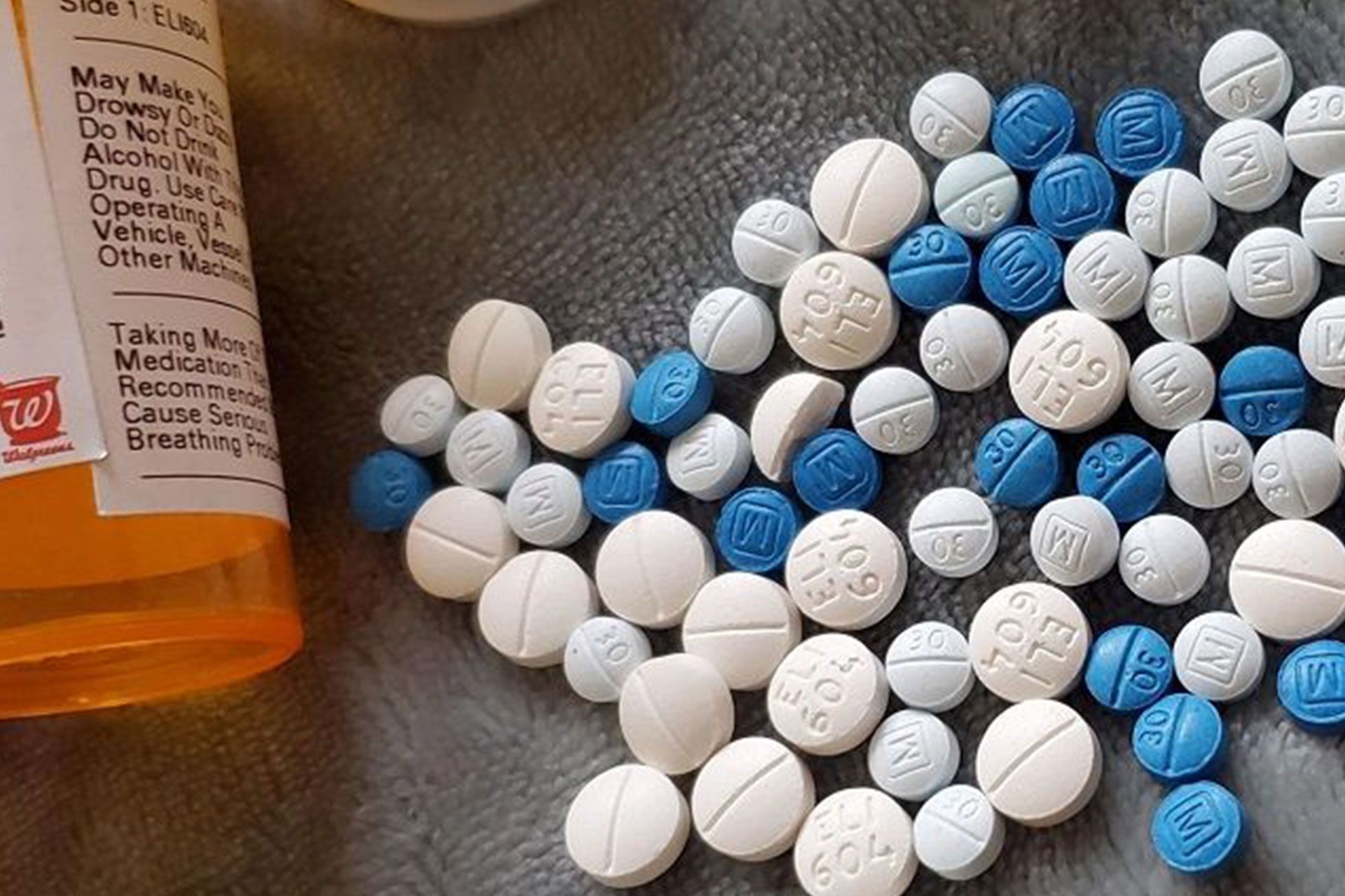 Fentanyl pills in various shades of white and blue scattered on a gray surface next to a prescription bottle.