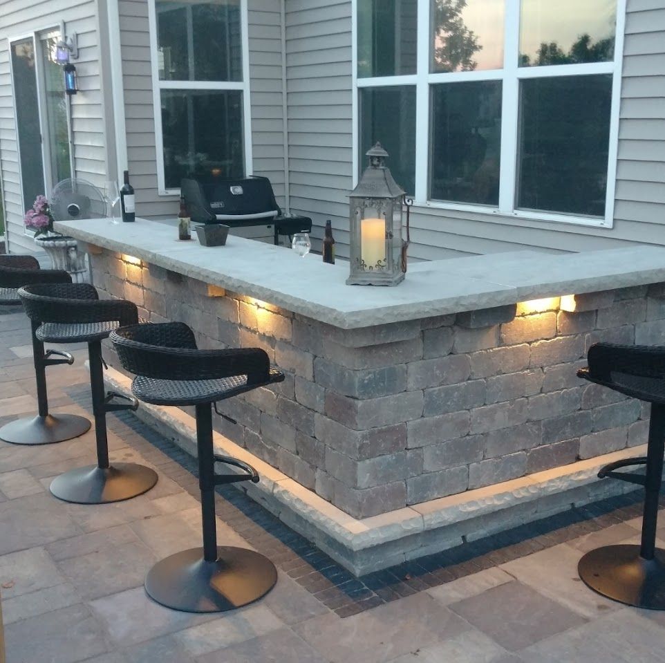 An outdoor stone bar on a patio, featuring four black stools, integrated warm lighting, and a decorative candle lantern.