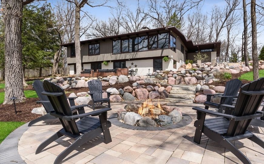A fire pit with lit flames sits on a stone patio surrounded by four black Adirondack chairs in front of a modern home.