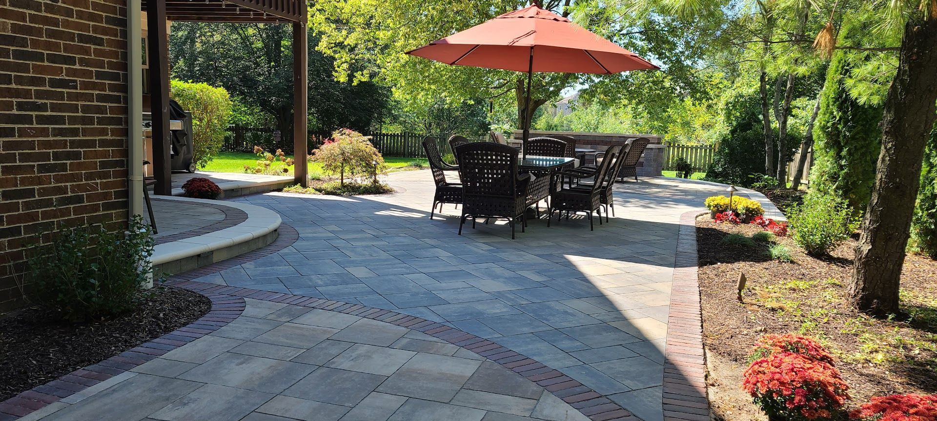 A sunny patio features gray patterned pavers, a reddish-orange umbrella, outdoor seating, and surrounding landscaping.