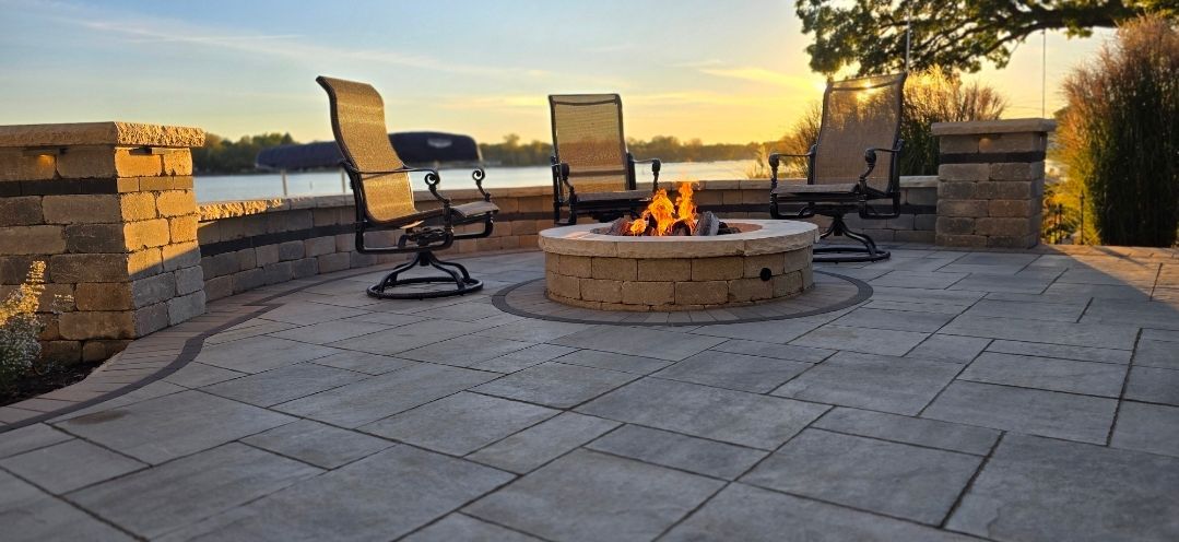 A lit stone fire pit on a paver patio with three outdoor chairs overlooking a lake at sunset.