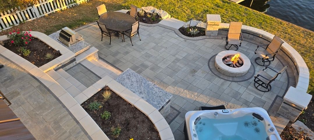 A stone patio featuring a fire pit, hot tub, outdoor dining area, and tiered garden beds near a waterfront.