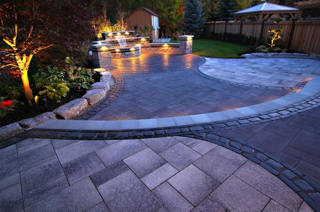 A stone patio at dusk with integrated landscape lighting, a water feature, and a backyard gazebo.