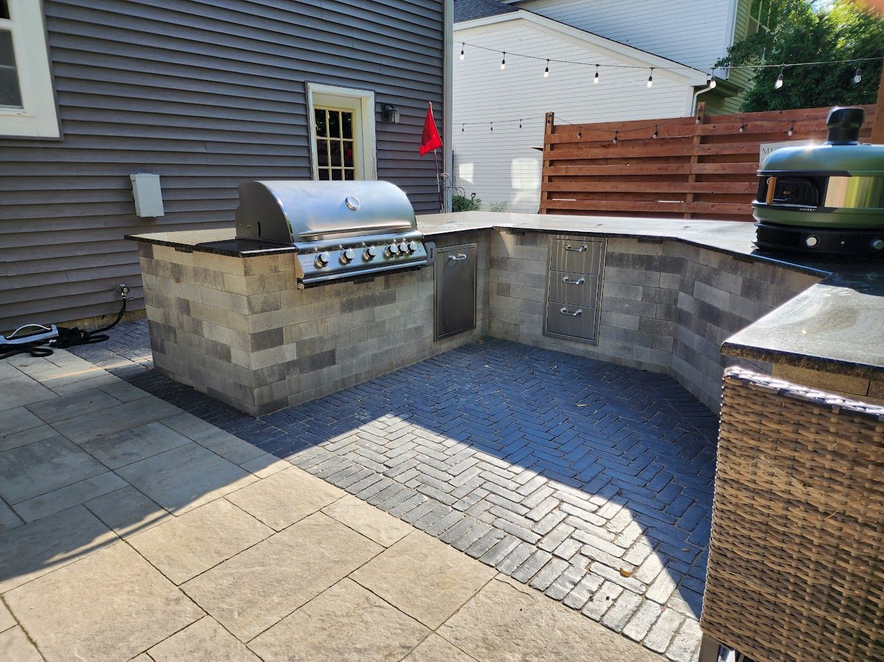 An outdoor kitchen with a stainless steel grill and pizza oven set on a stone patio against a gray house.