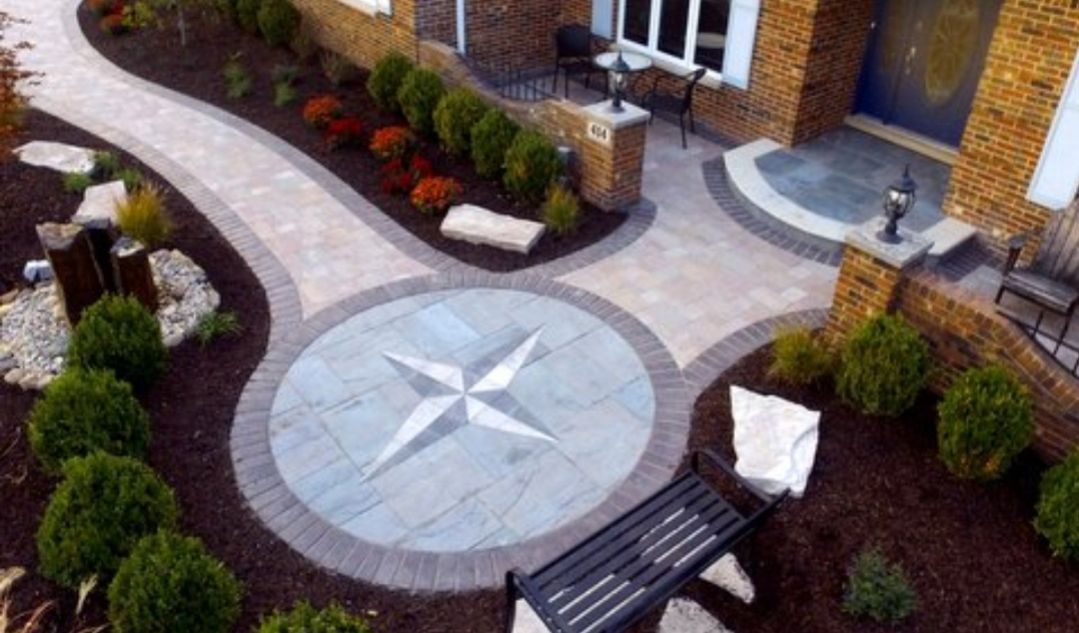 A stone patio featuring a decorative star inlay leads to a house entrance, surrounded by garden beds and a black bench.