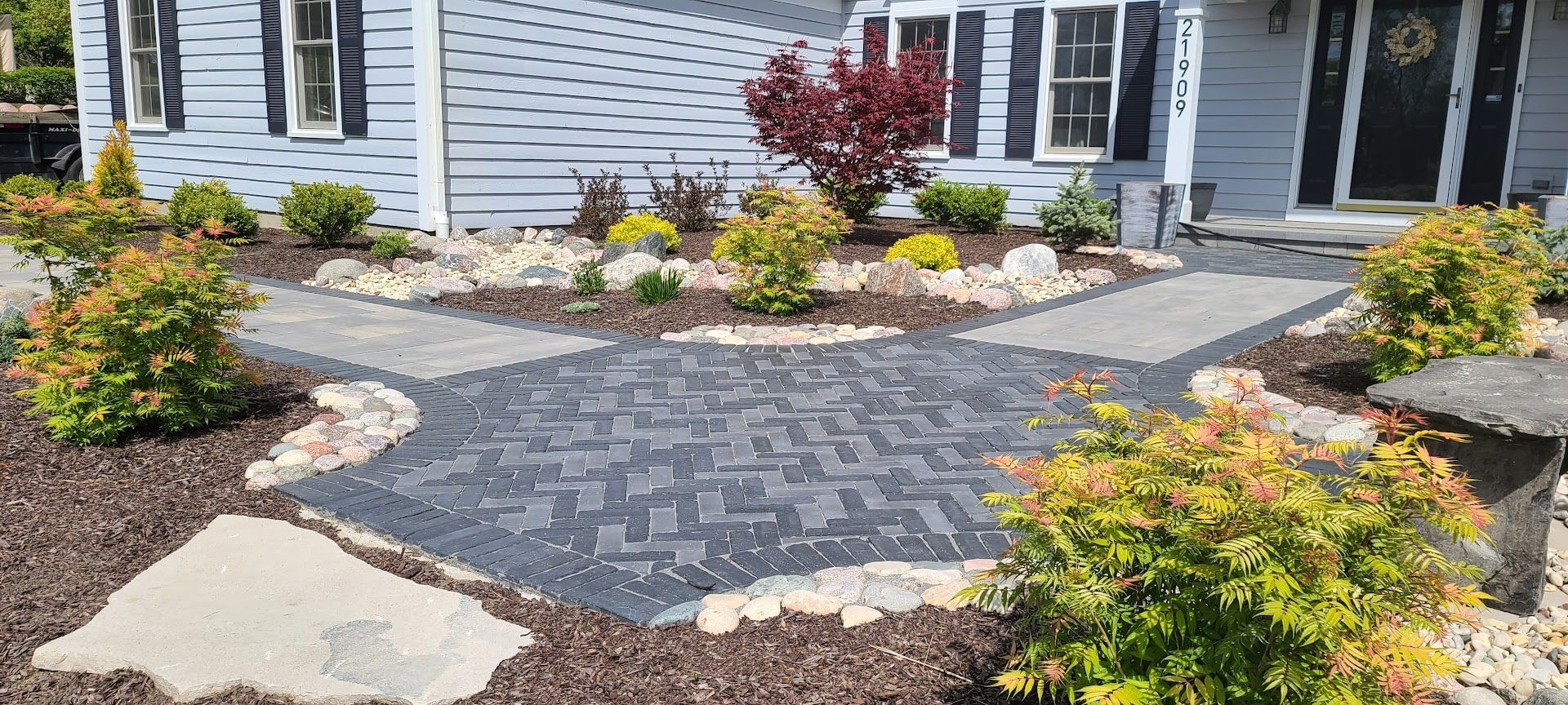 A landscaped front yard featuring a herringbone-patterned paver walkway leading to a light blue house entrance.