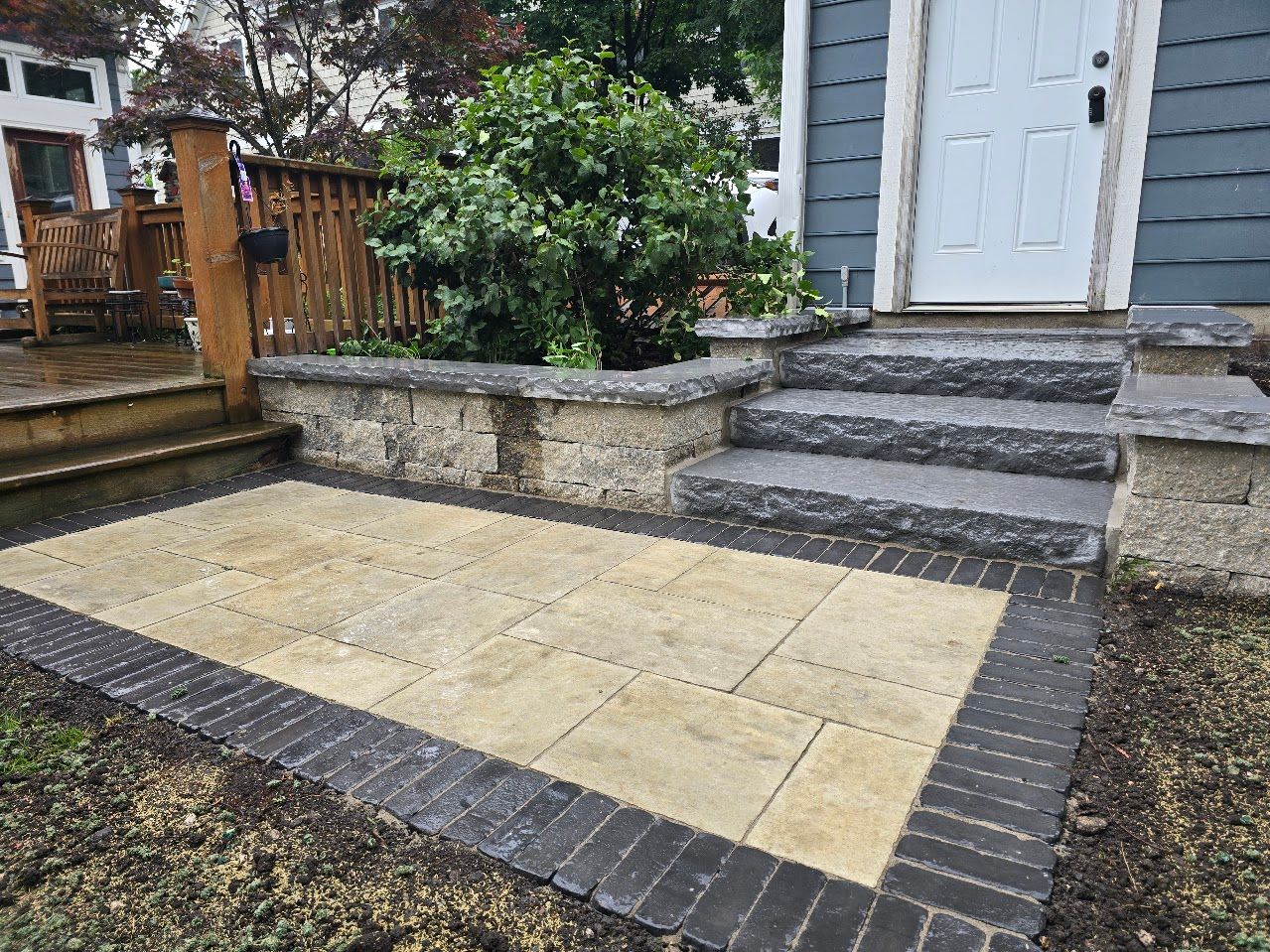 A patio with tan rectangular pavers, bordered by dark bricks, leading to stone steps and a retaining wall by a house.
