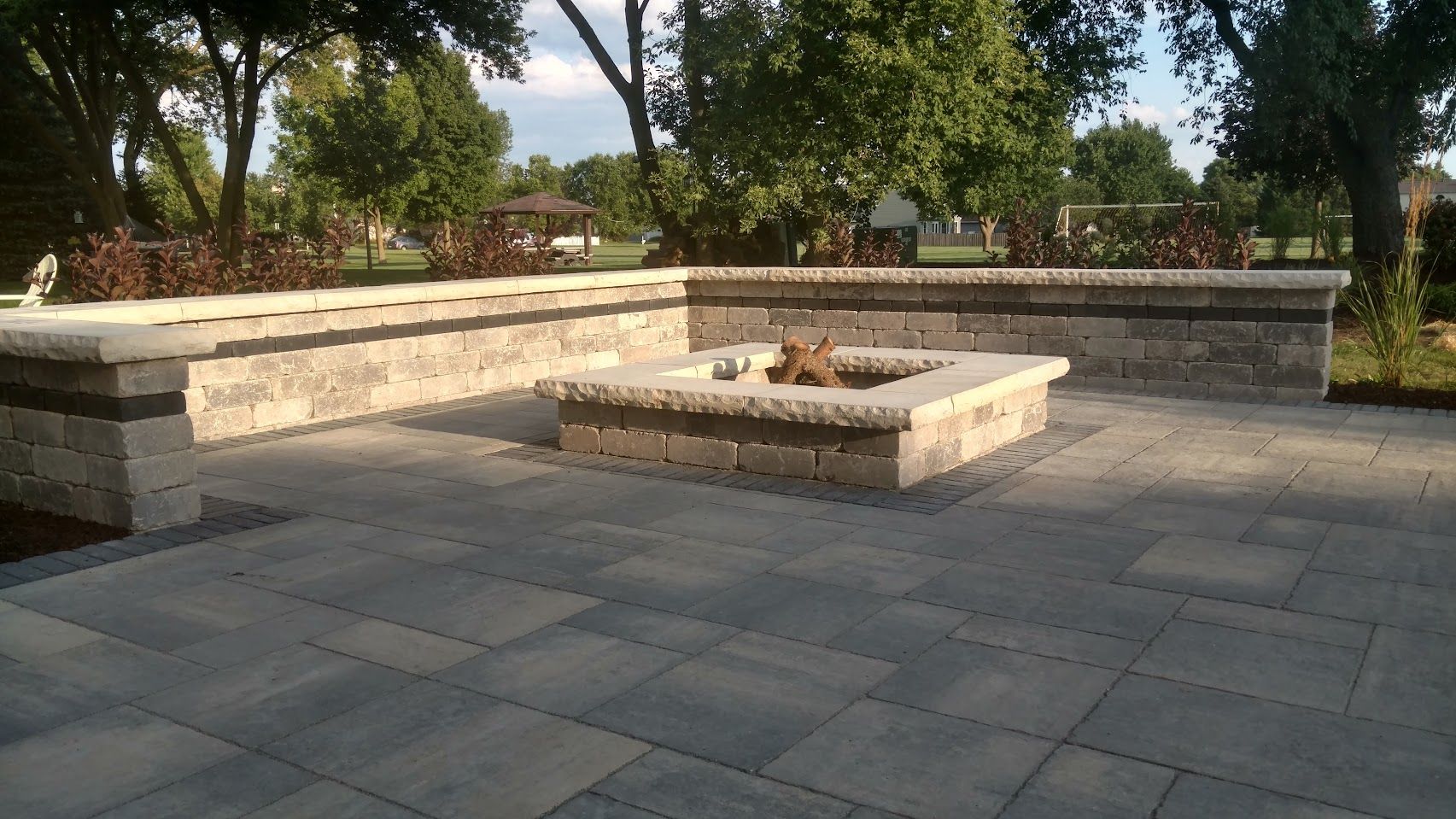 A stone patio featuring a square fire pit and a low, L-shaped stone retaining wall seating area, surrounded by trees.