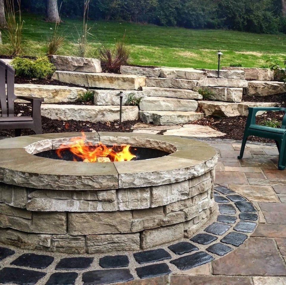 A circular stone fire pit with a roaring fire on a patio, set before terraced stone retaining walls and a green lawn.