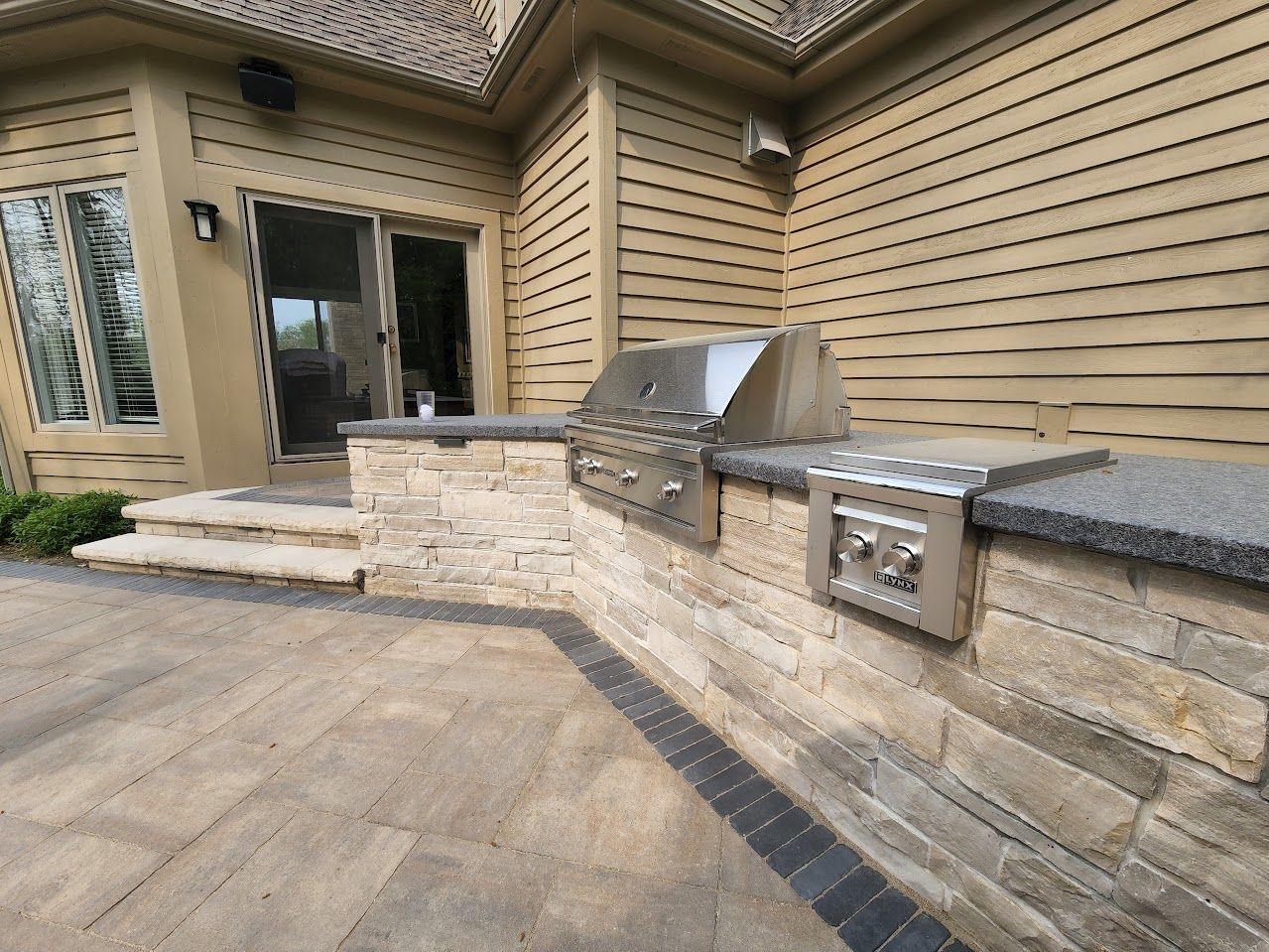 An outdoor kitchen featuring a stainless steel grill and side burner set into a light-colored stone wall on a stone patio.