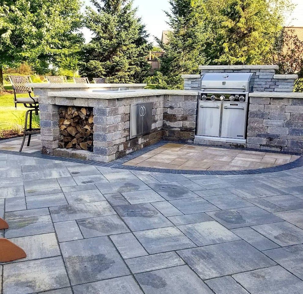 Outdoor stone kitchen with a built-in grill, firewood storage, and bar seating on a paved patio in a grassy backyard.