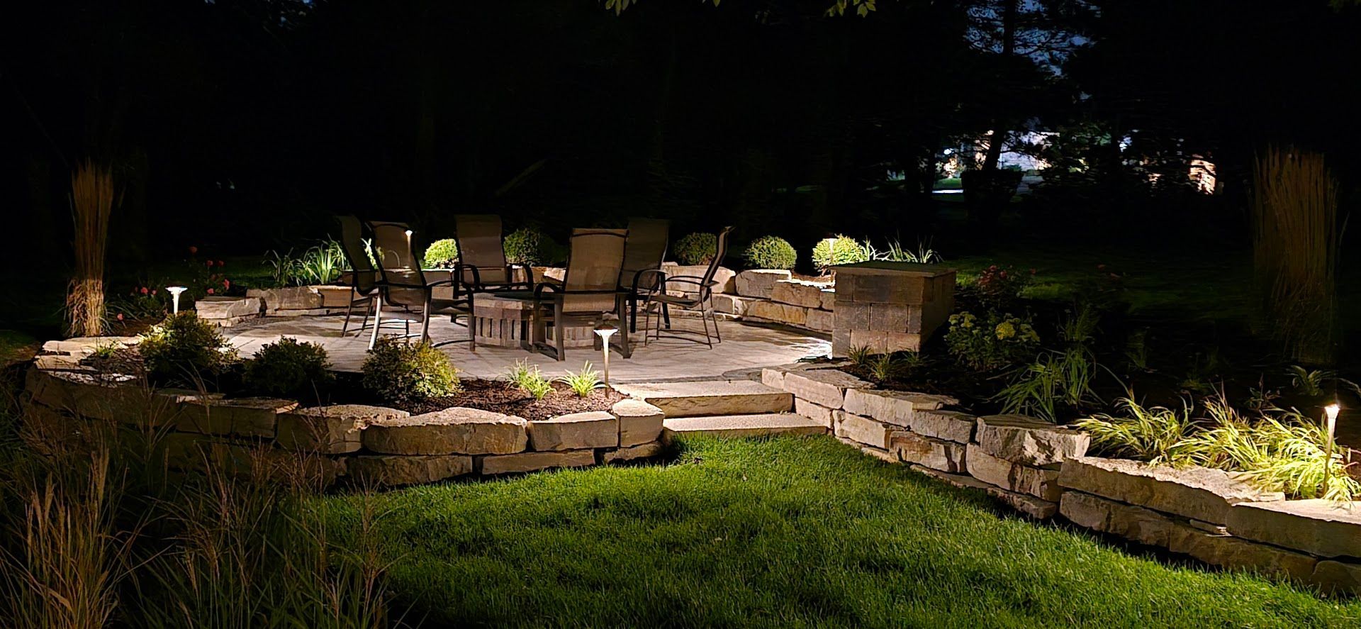A stone patio with outdoor furniture is illuminated at night, surrounded by landscaped garden beds and low-voltage lighting.