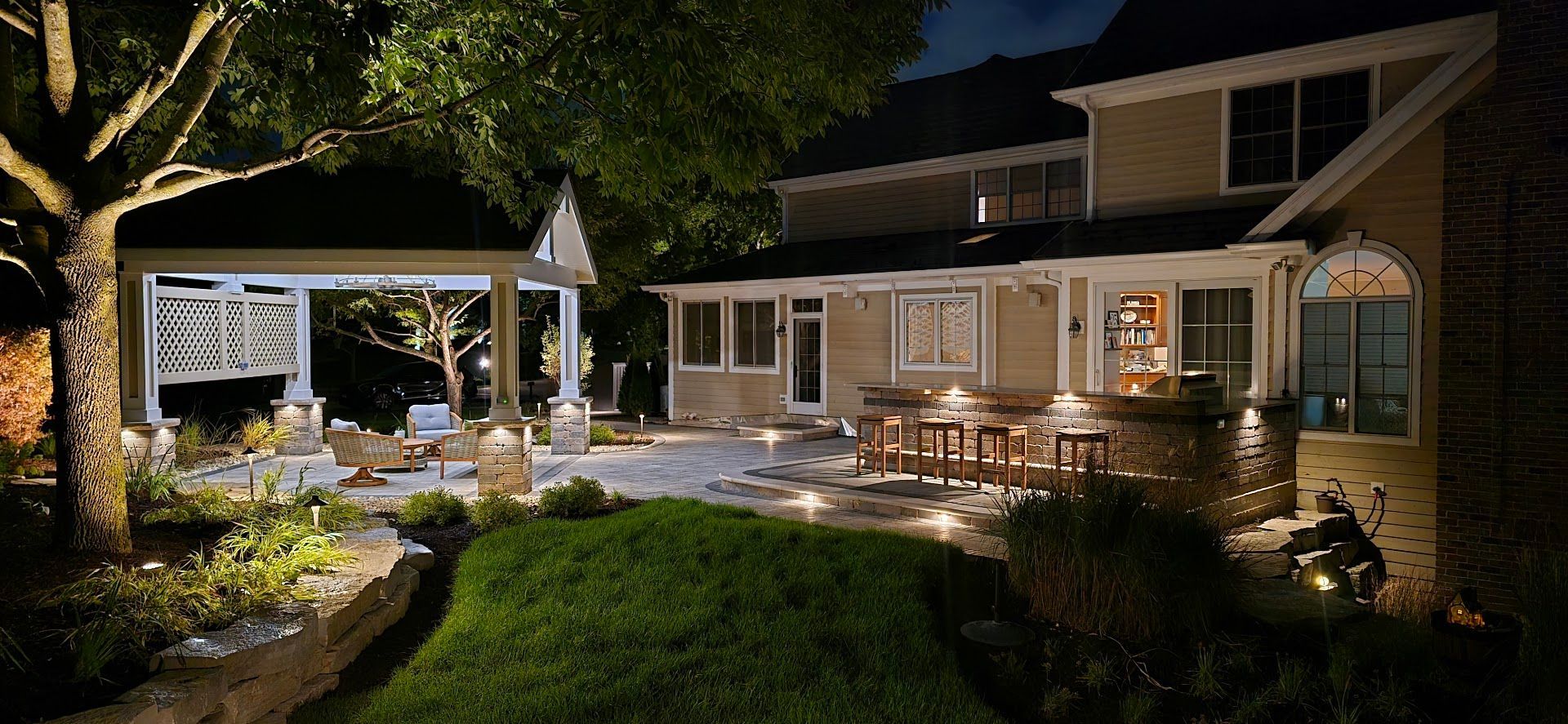 An illuminated backyard patio at night featuring a stone bar, seating area, gazebo, and manicured landscaping.