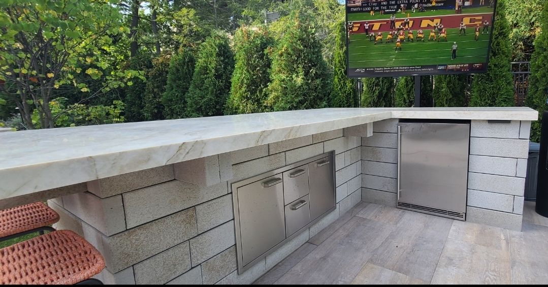 Outdoor kitchen with a stone counter, stainless steel storage, and a built-in refrigerator under a mounted TV.