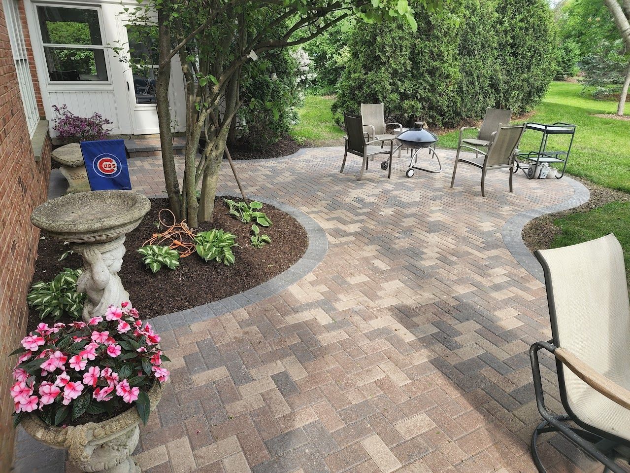 A patio with multi-toned stone pavers, a tree in a landscaped bed, a stone birdbath, and outdoor seating.