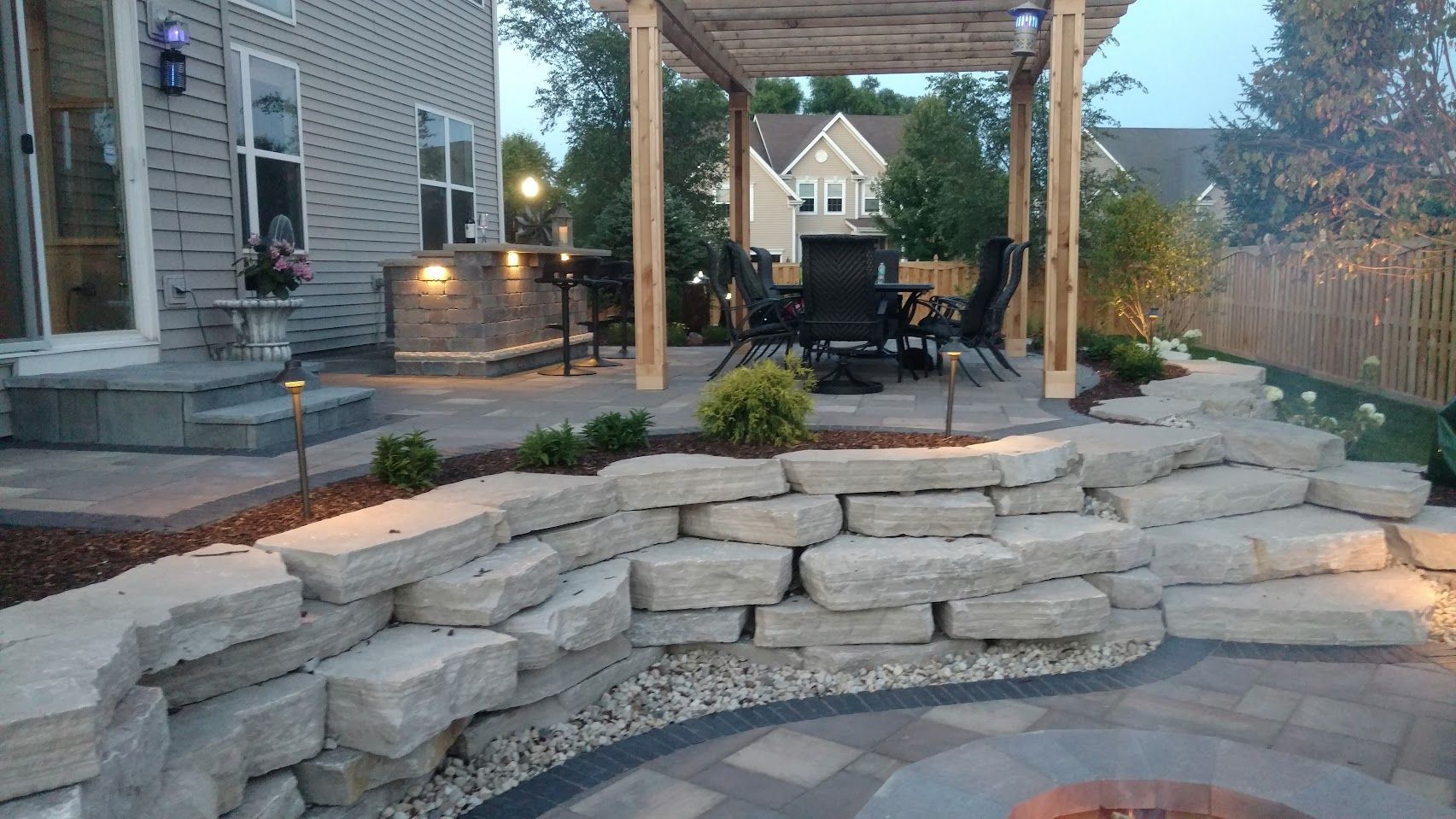 A multi-level backyard patio features a stone retaining wall, outdoor kitchen, pergola-covered dining area, and fire pit.