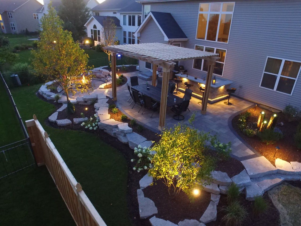 Aerial view of a patio at night featuring a pergola, dining area, outdoor kitchen, and landscaped lighting in a backyard.